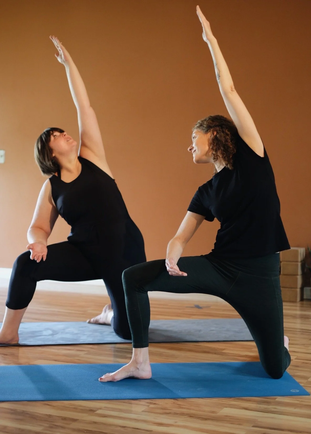 Two women practicing yoga indoors on mats, both in a lunge pose with one arm stretched upward and smiling at each other.