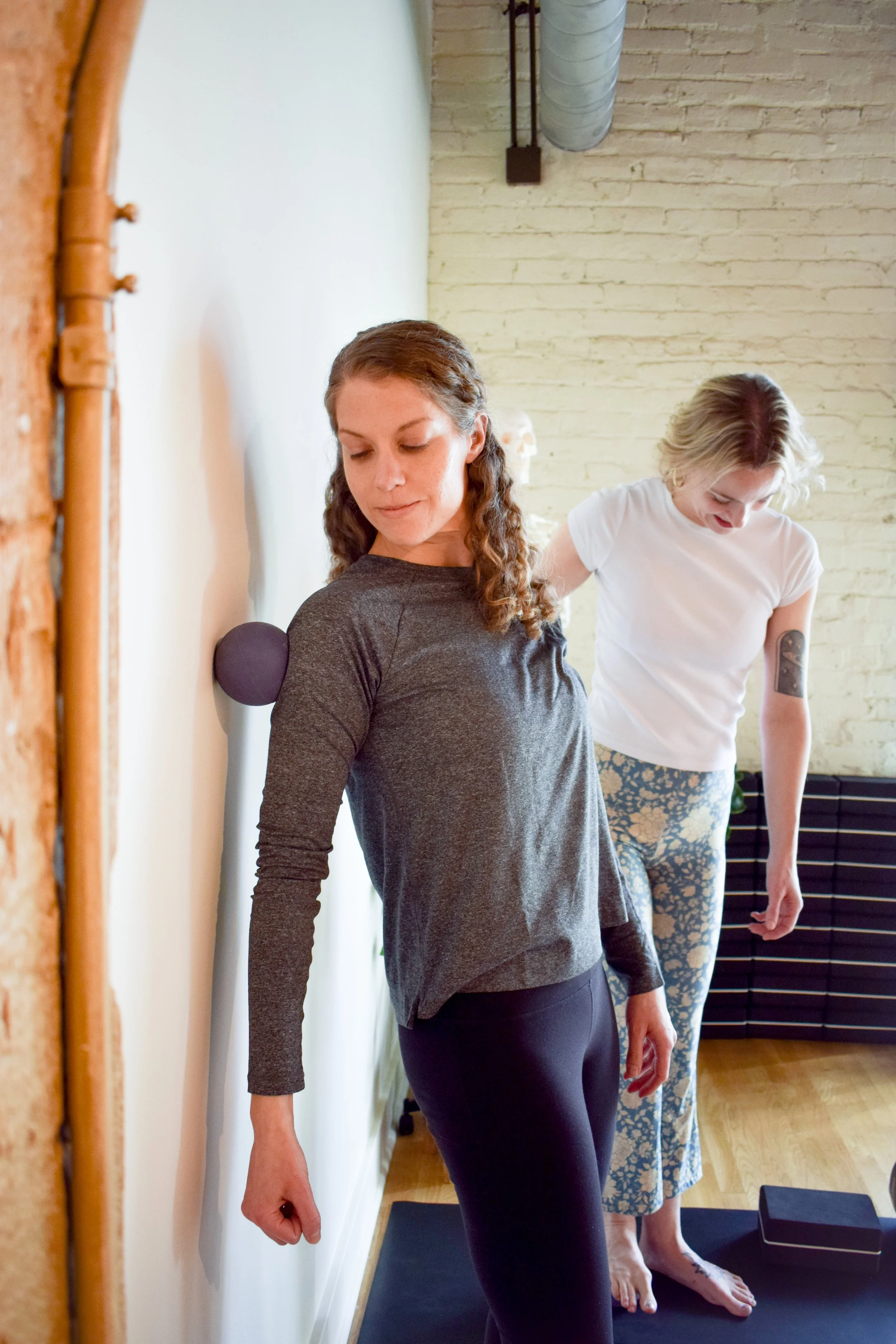 Two women are practicing yoga, one leaning against a wall with a purple massage ball on her shoulder, the other standing on a yoga mat wearing patterned leggings, in a room with exposed brick and ductwork.
