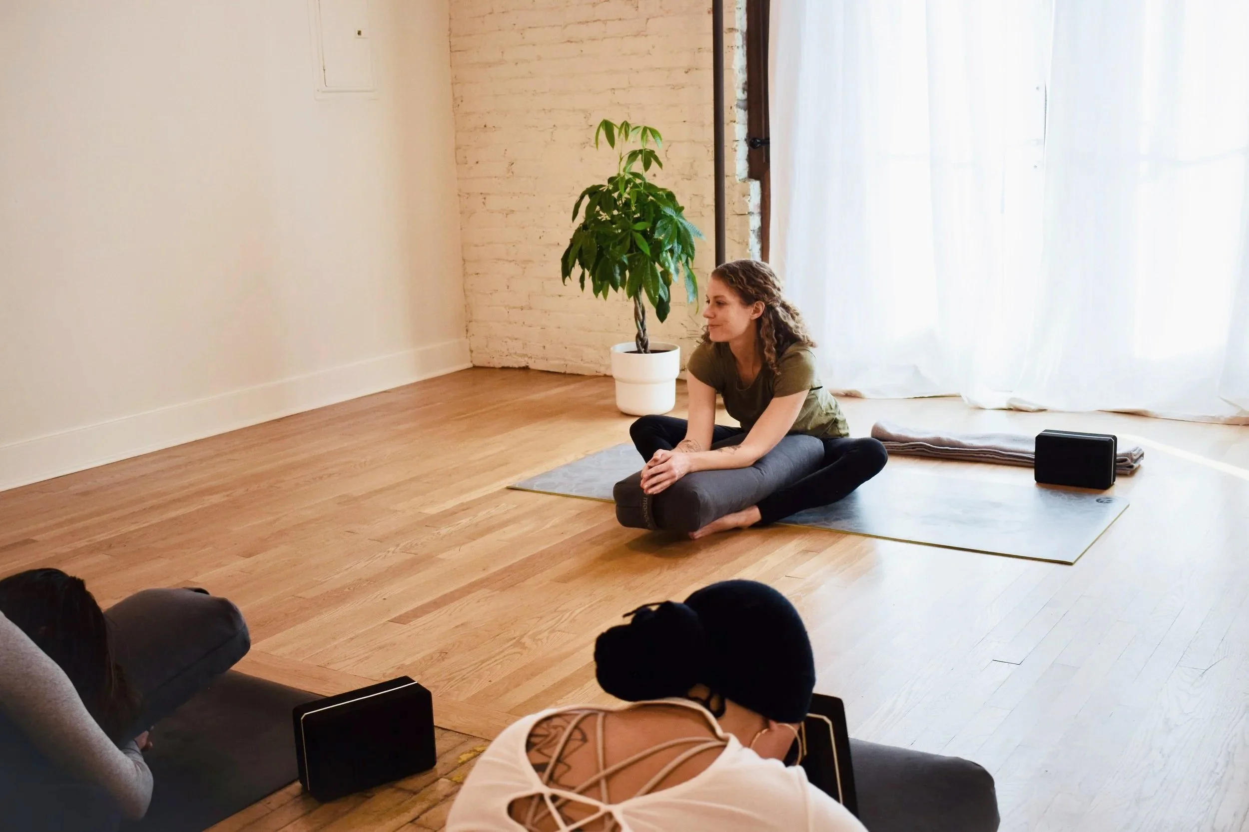 A woman sitting on a yoga mat in a room with wooden floors and white walls, facing two people lying on the floor with yoga blocks. There is a green potted plant and white curtains in the background.