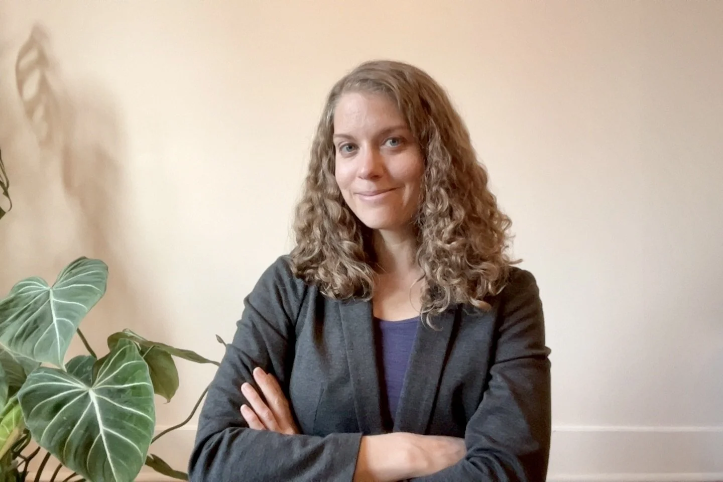 A woman with curly blonde hair, wearing a dark blazer, standing with arms crossed in front of a beige wall, with a large green leafy plant on her left.