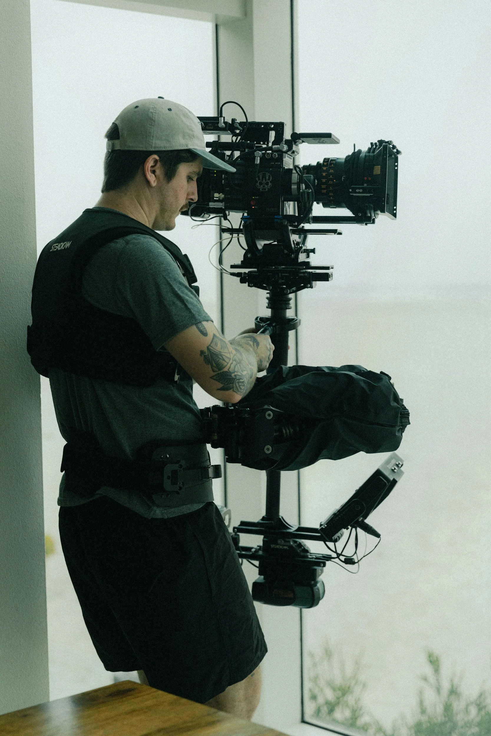 A person wearing a gray cap, black vest, and black shorts operating a professional film camera on a steadicam rig by a large window.