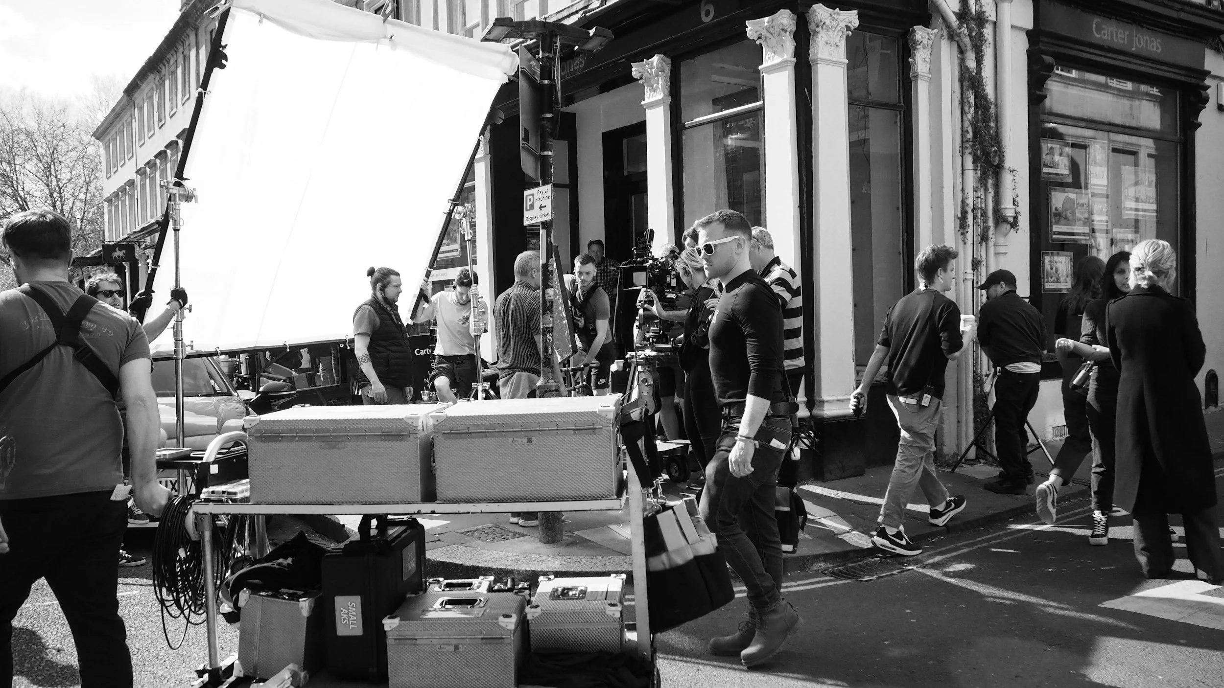 Film crew working on a street outside a storefront with several people walking by, and filming equipment set up including lights, cameras, and cases.