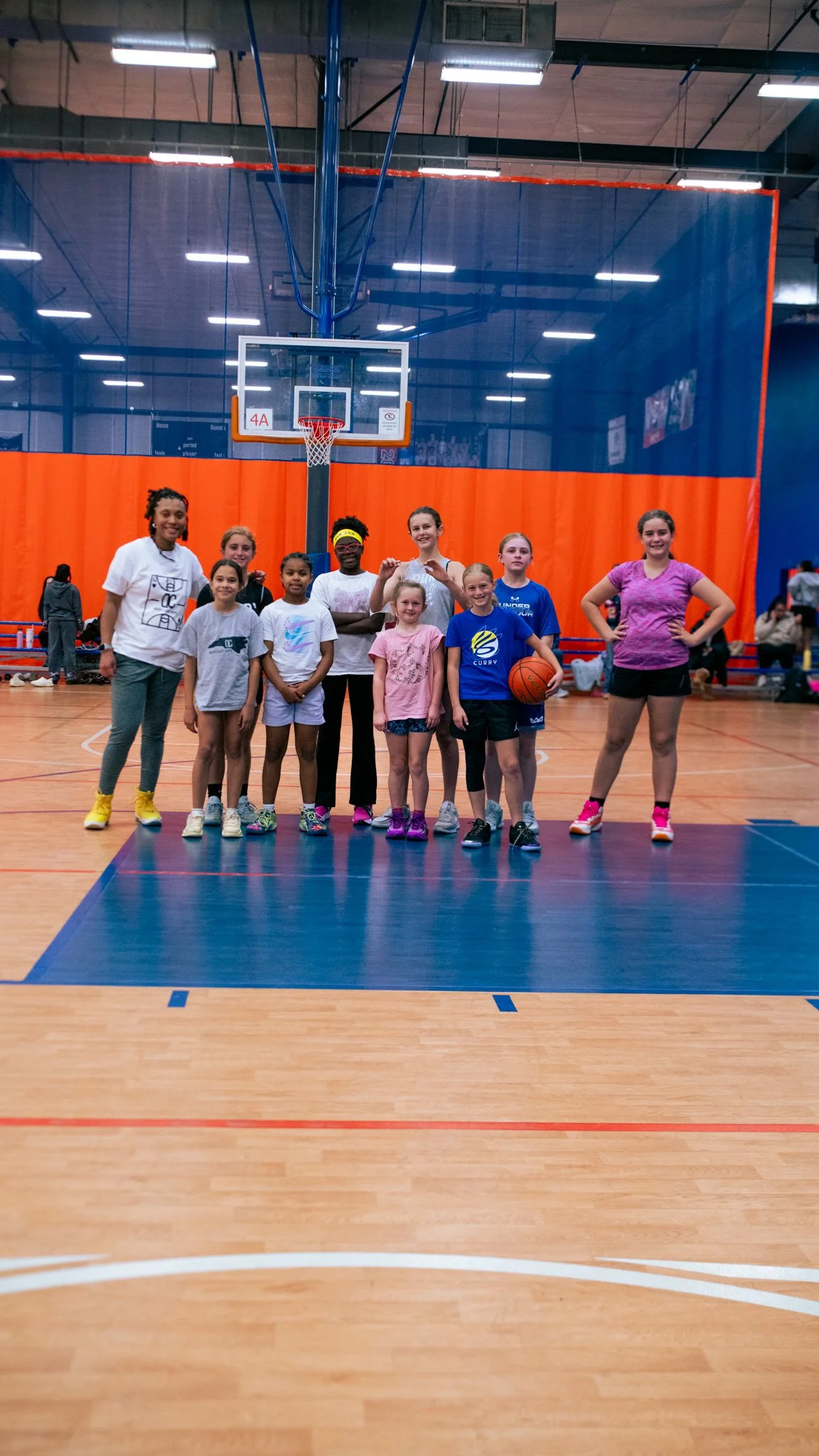 A group of children and teenagers in a gymnasium, standing in front of a basketball hoop, posing for a photo, some holding basketballs.