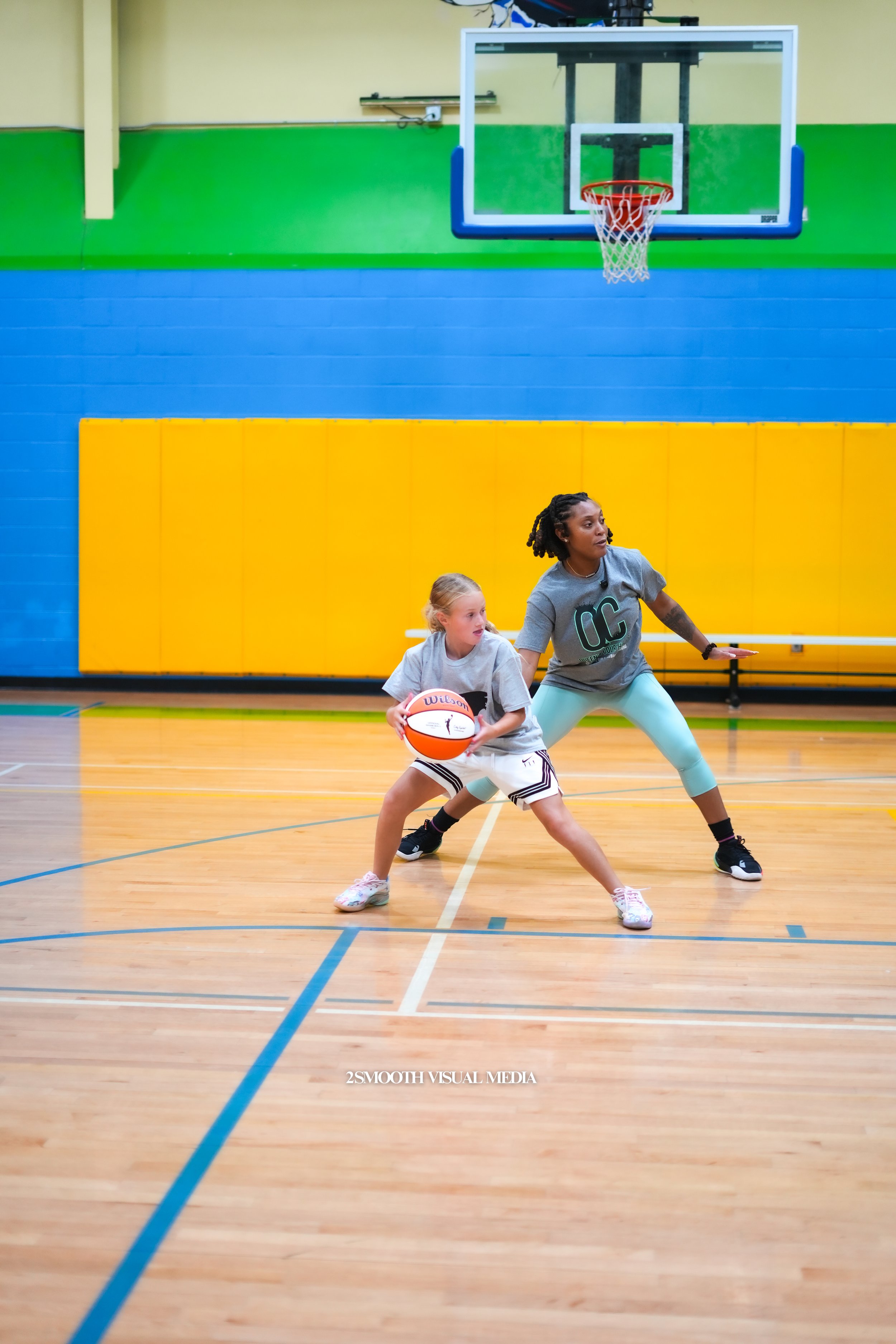 A young girl holding a basketball on a basketball court with a woman practicing guard stance behind her, inside a gym with yellow and green walls and a basketball hoop overhead.