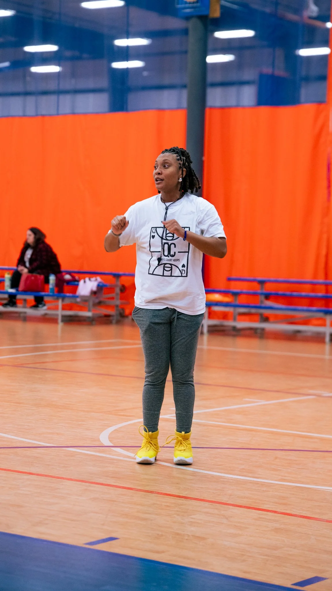 A woman in a white T-shirt, gray leggings, and yellow sneakers standing on an indoor basketball court, with orange and blue seats and a woman sitting in the background.