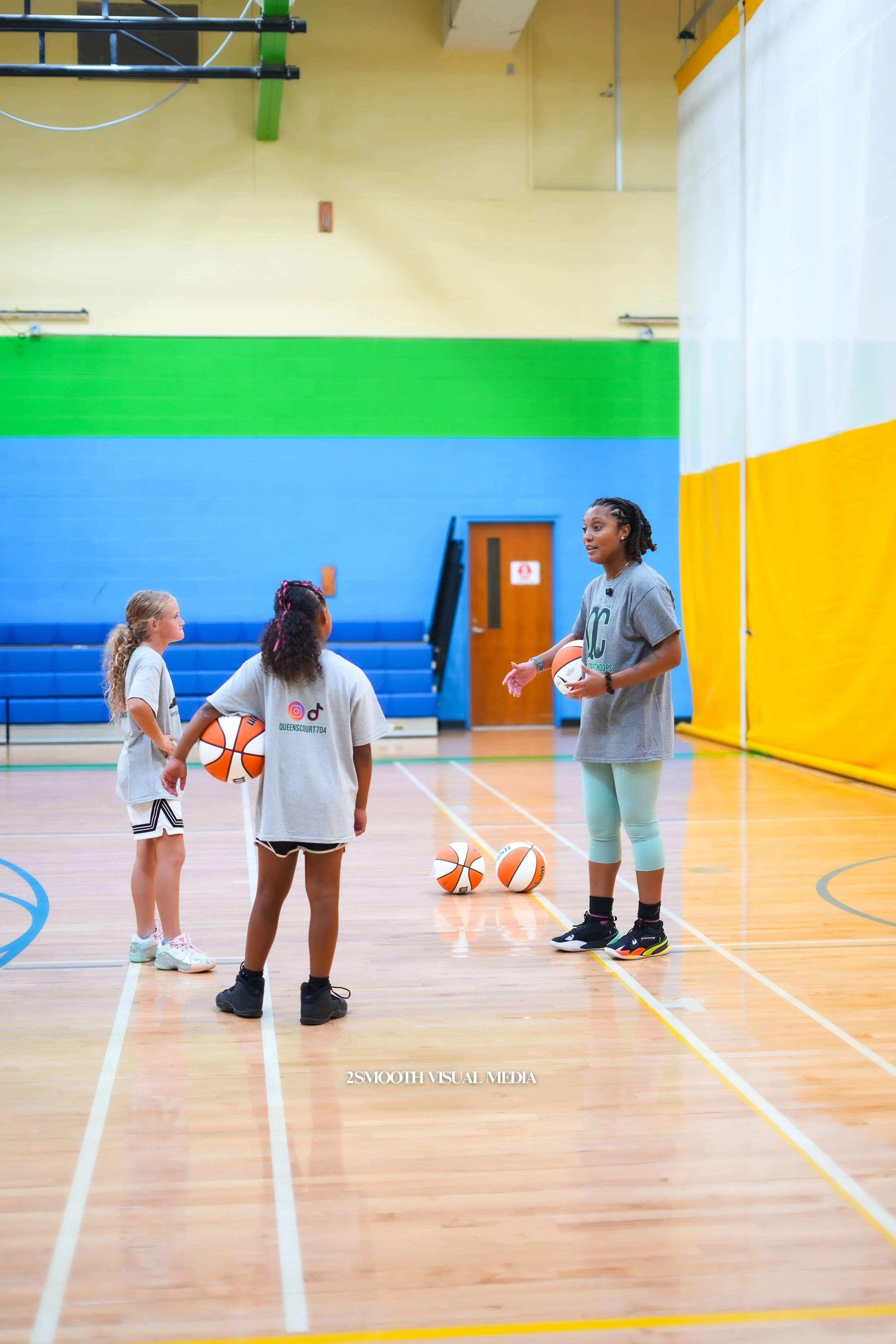 A female basketball coach giving instructions to two young girls holding basketballs in an indoor gymnasium, with a yellow, green, and blue wall in the background.