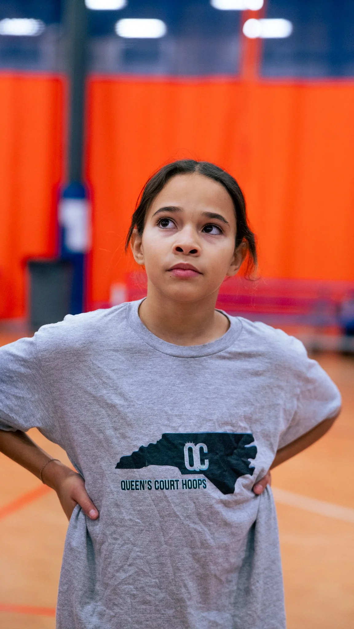 A young girl standing with her hands on her hips, wearing a gray T-shirt with a silhouette of North Carolina and the words "Queen's Court Hoops" printed on it, inside a gymnasium with orange walls and a basketball hoop in the background.