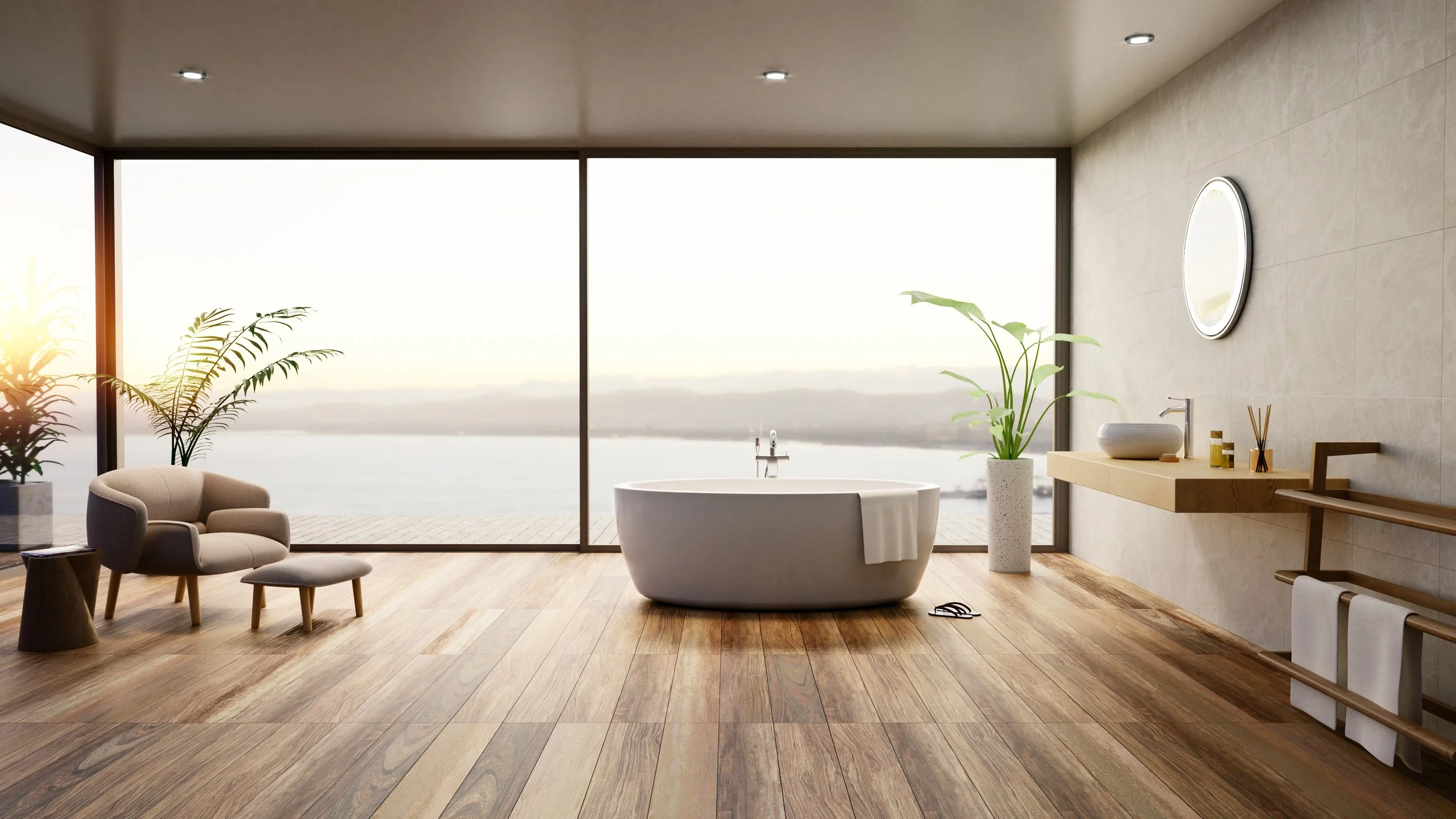 A modern bathroom with a freestanding bathtub in front of large floor-to-ceiling windows overlooking a body of water. The room has wooden flooring, a beige wall with a round mirror, a wooden vanity with a vessel sink, potted plants, and minimal decor.