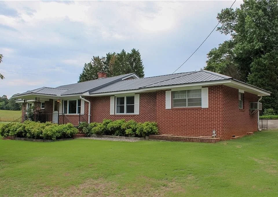 A single-story brick house with a gray metal roof, surrounded by a well-maintained lawn and bushes. There are trees in the background and a cloudy sky overhead.