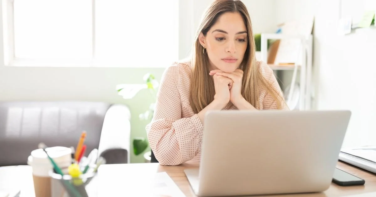 A woman sitting at a desk, looking at a laptop with a thoughtful expression, in a bright room with large windows and houseplants.