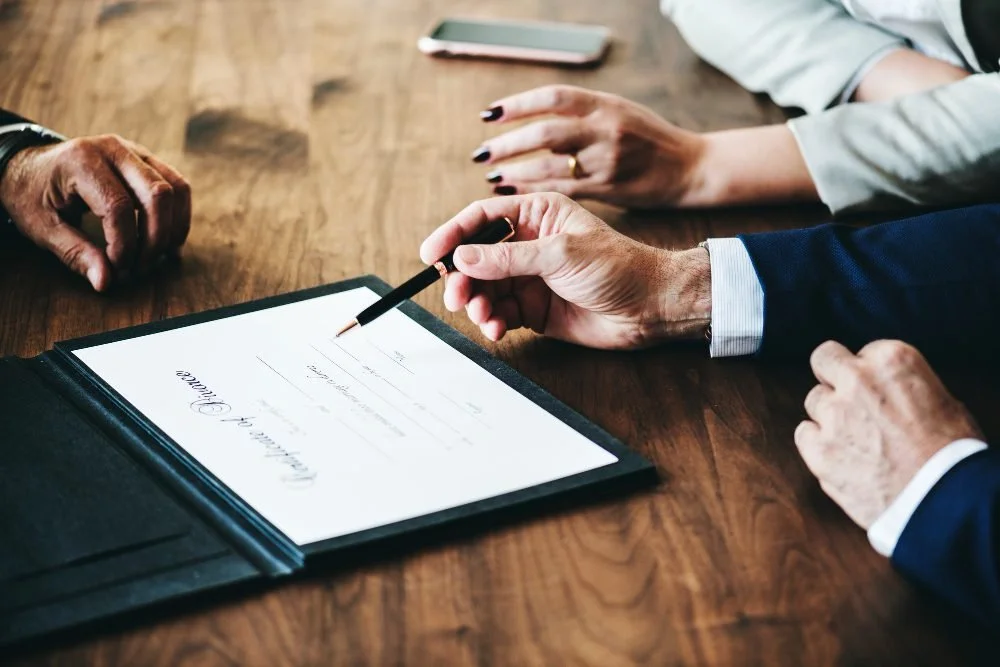 Two professionals signing a contract or legal document on a wooden table, with a tablet and a smartphone nearby.