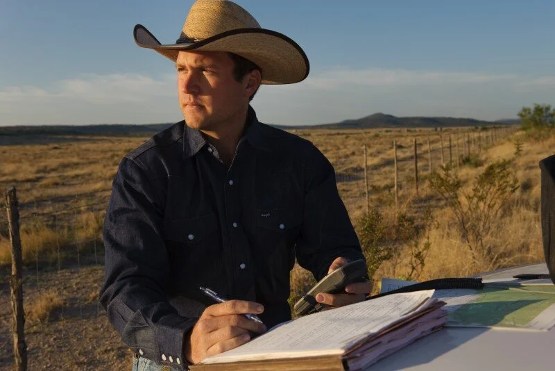 A man wearing a cowboy hat and dark shirt standing outdoors in a rural landscape, holding a phone in one hand and a pen in the other, with open notebooks and papers on a surface in front of him.