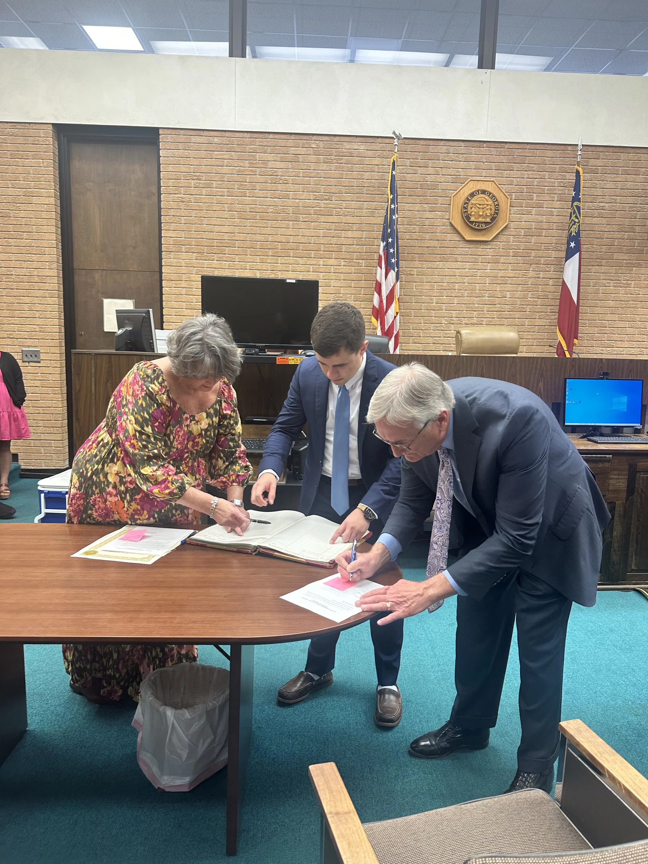 Three people, two men and one woman, signing documents at a wooden table in a formal setting, with an American flag and a state flag behind them, and a brick wall with a round official seal on it.
