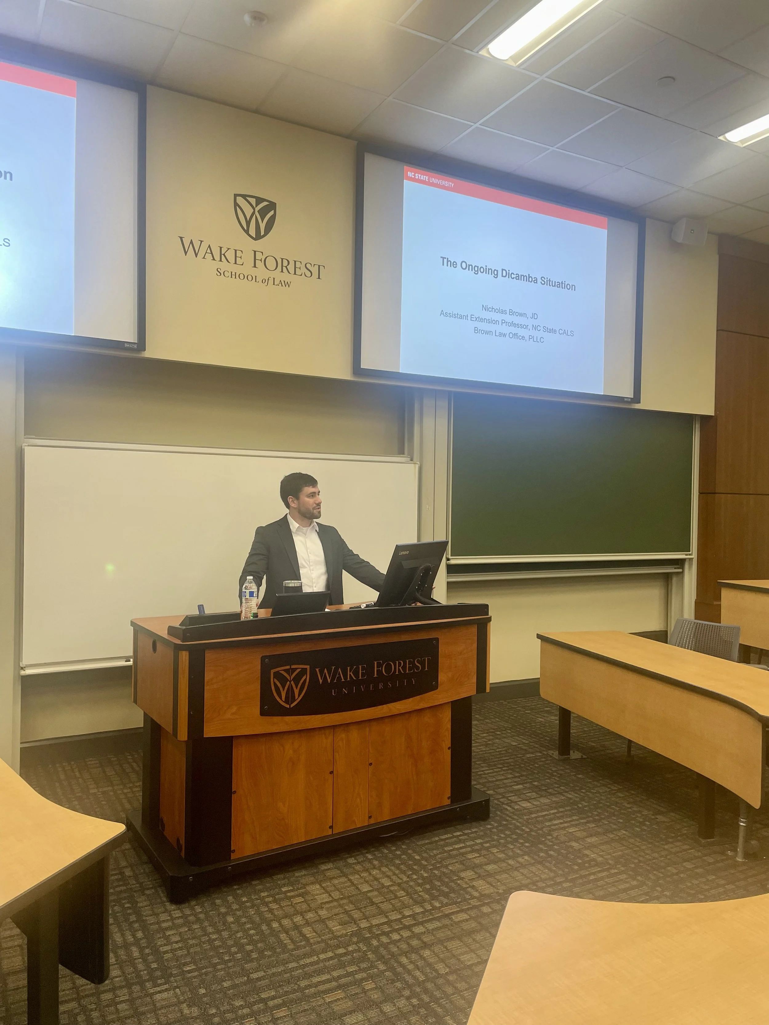 A man standing at a podium giving a presentation in a classroom at Wake Forest University School of Law. The presentation slide reads 'The Ongoing Dicamba Situation.'