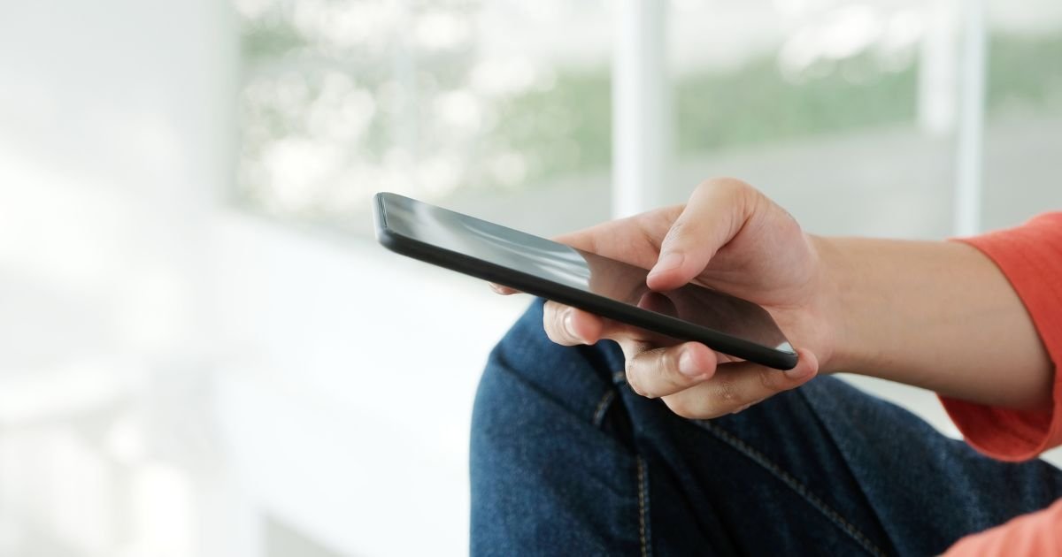 Person holding a smartphone while sitting with a blurred background of windows and greenery.