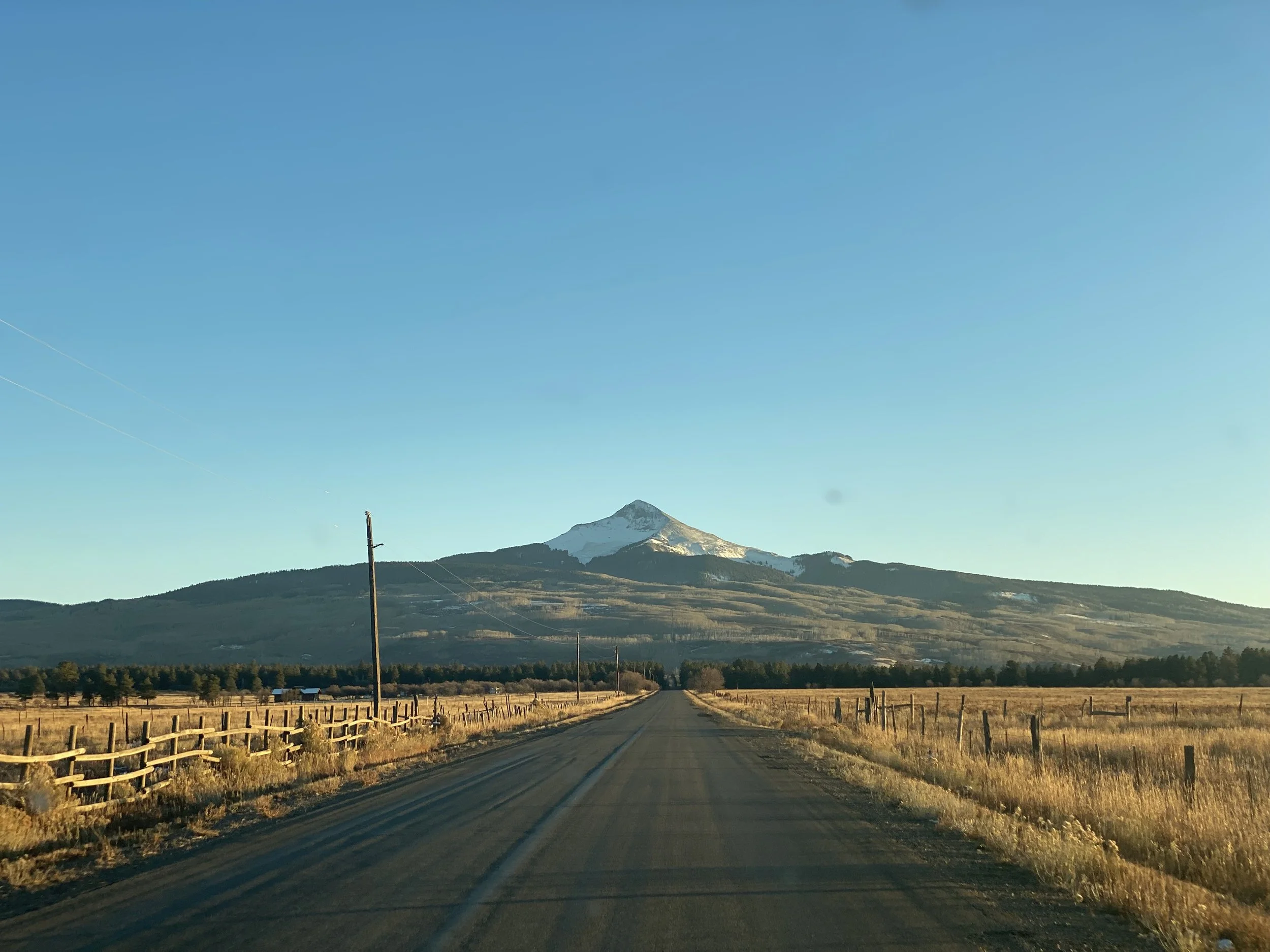 A long rural road leading towards a mountain with a snow-capped peak under a clear blue sky, flanked by fields with wooden fences.