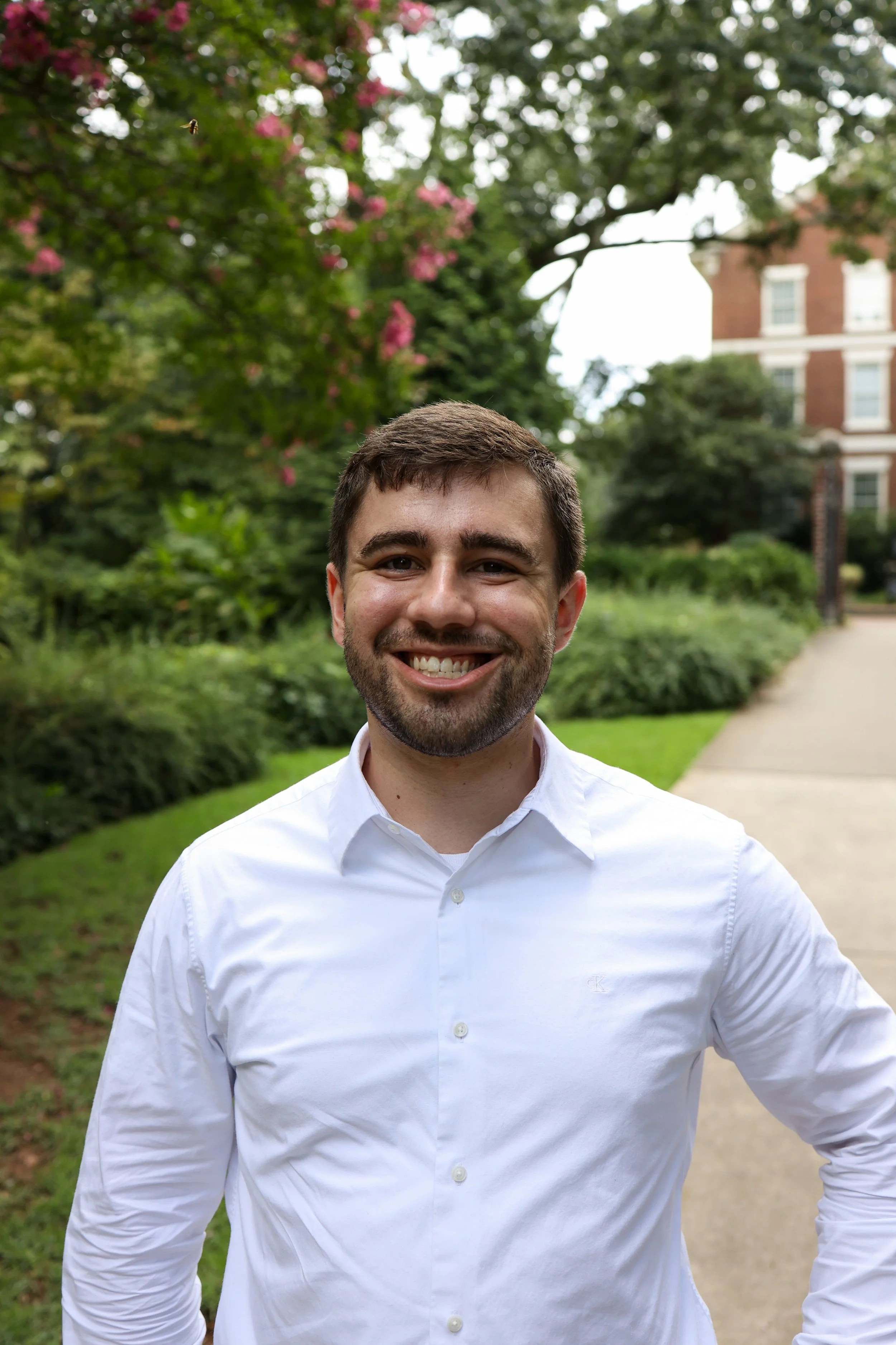 A smiling man with dark brown hair, beard, and light skin, dressed in a white button-down shirt, standing outdoors on a sidewalk with greenery and a building in the background.