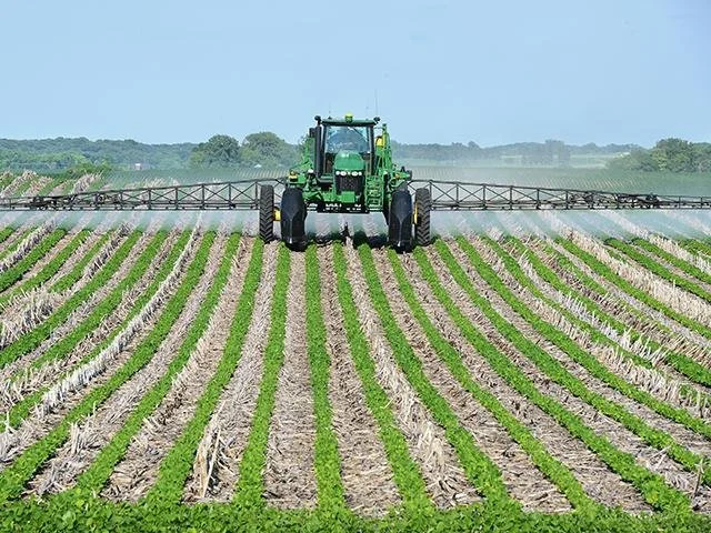 A green tractor spraying crops in a large farm field with rows of young plants.