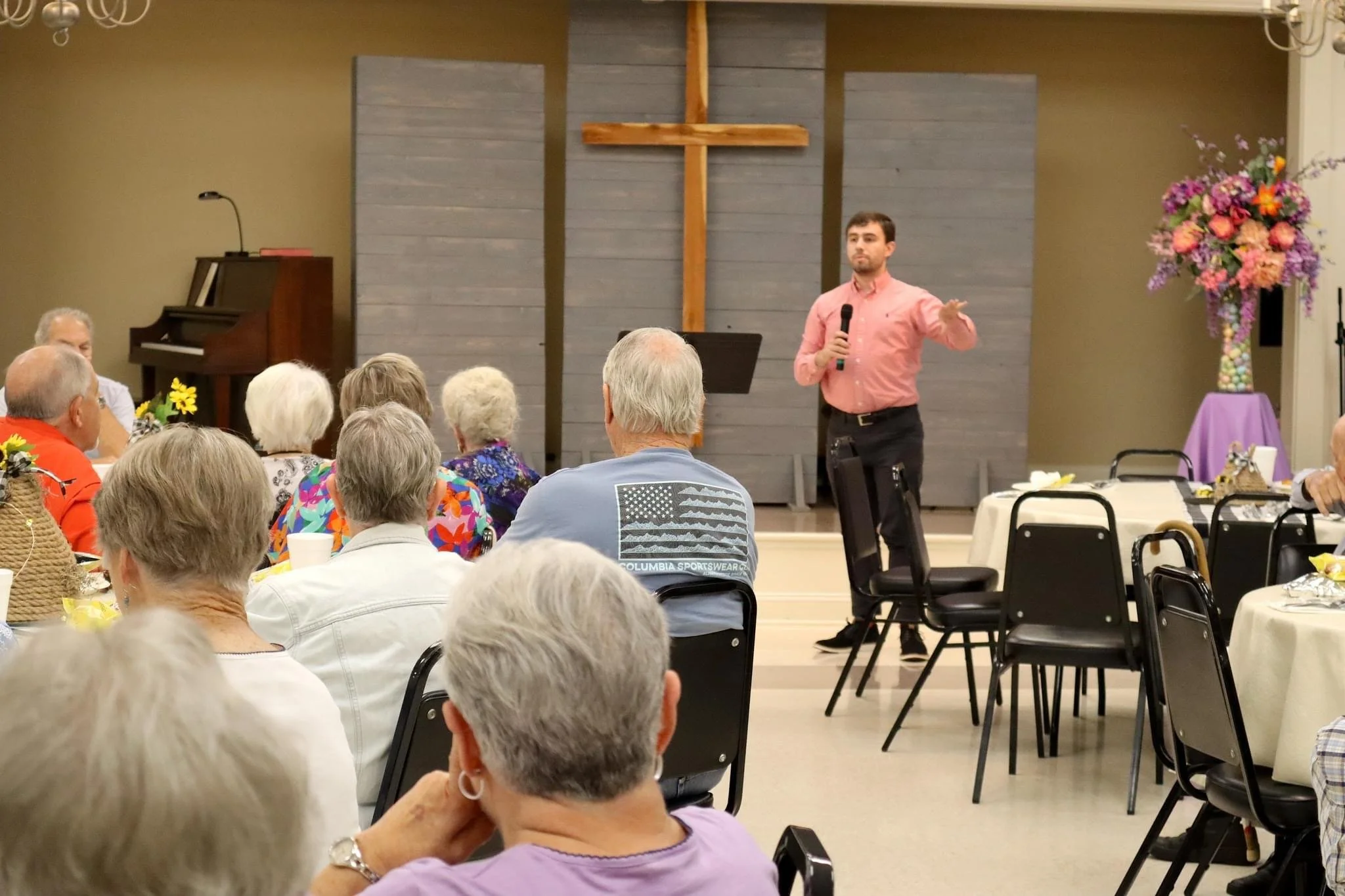 A man in a pink shirt holding a microphone and gesturing while speaking to a seated audience in a church or community hall. The background features a cross on the wall and a flower arrangement on the right.