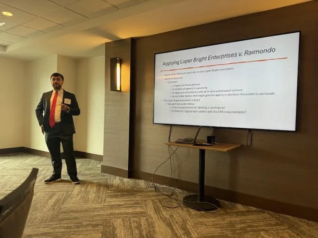 A man in a dark suit, white shirt, and red tie stands next to a large wall-mounted screen displaying a presentation slide in a conference room. The slide is titled 'Applying Loper Bright Enterprises v. Raimondo' and includes bullet points. The room has beige walls, a carpeted floor, and a small table below the screen with electronic devices connected by visible cords.
