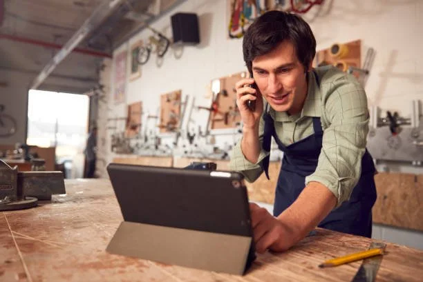 Man in a workshop talking on the phone and working on a laptop on a workbench.