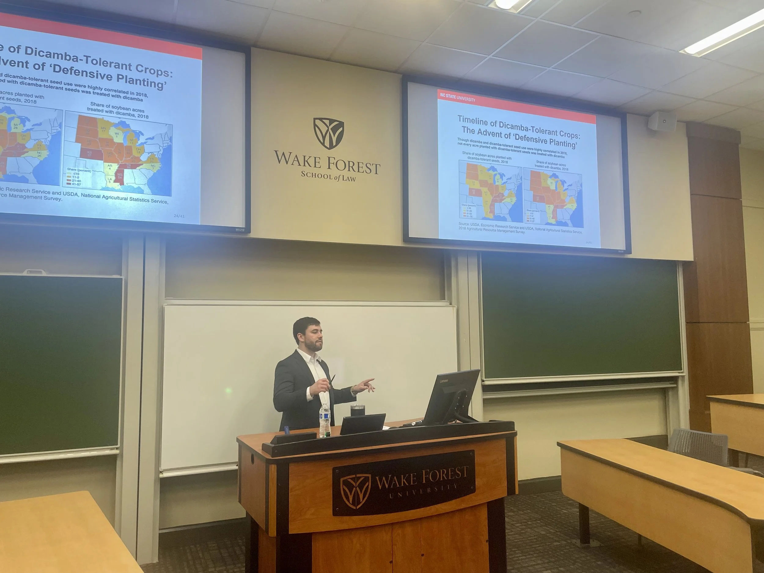 A man giving a presentation at Wake Forest University classroom, with two large screens displaying a slide titled 'Timeline of Dicamba-Tolerant Crops: The Advent of Defensive Planting', showing maps of the United States with data on soybean acres treated with dicamba in 2018.