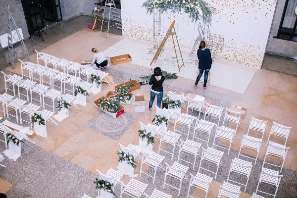 A large indoor space being prepared for an event, with multiple white chairs decorated with greenery, a flower arrangement, and three women arranging decorations on the chairs.