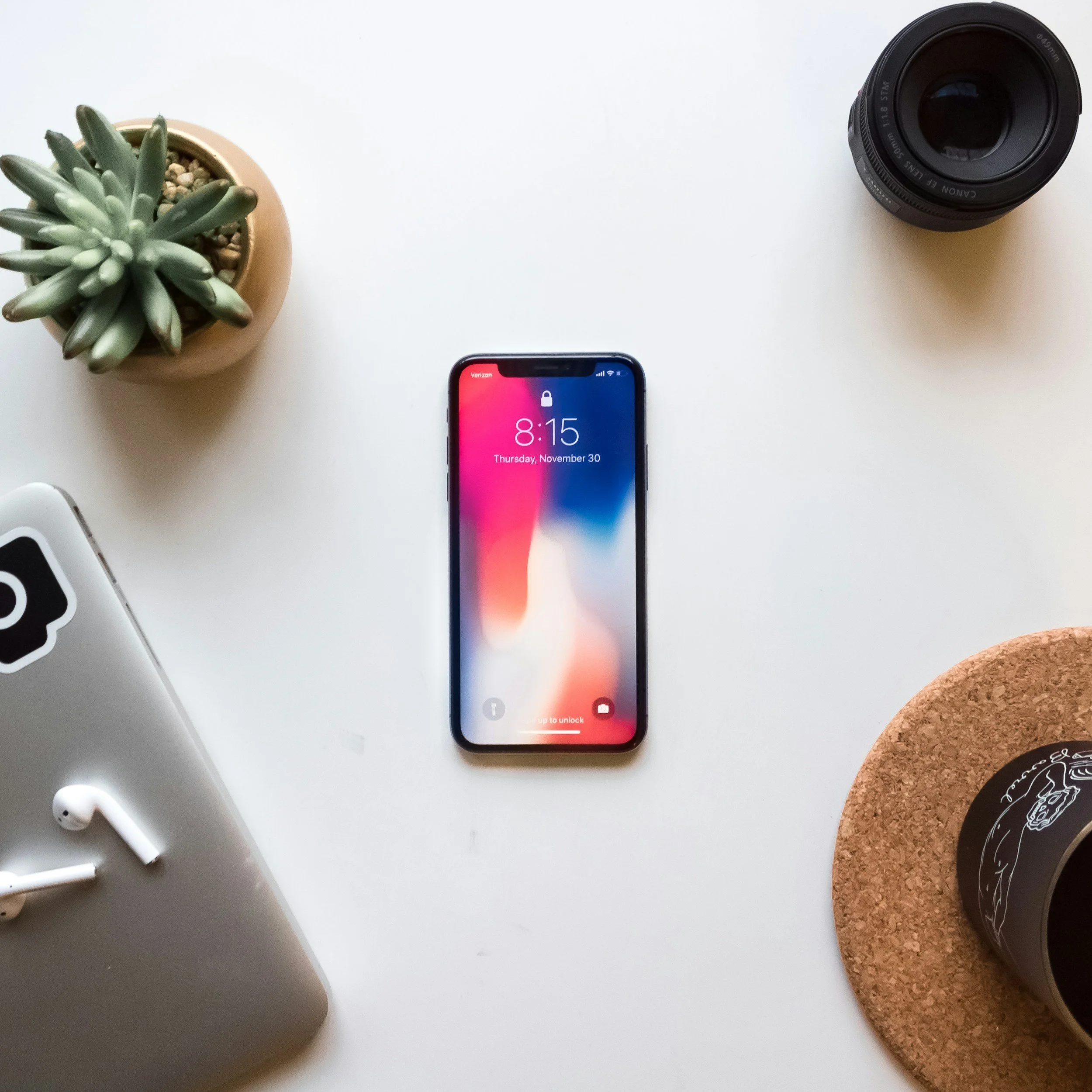 Top-down view of a white desk with an iPhone, a laptop with AirPods, a potted succulent, a camera lens, and a coffee cup on a cork coaster.