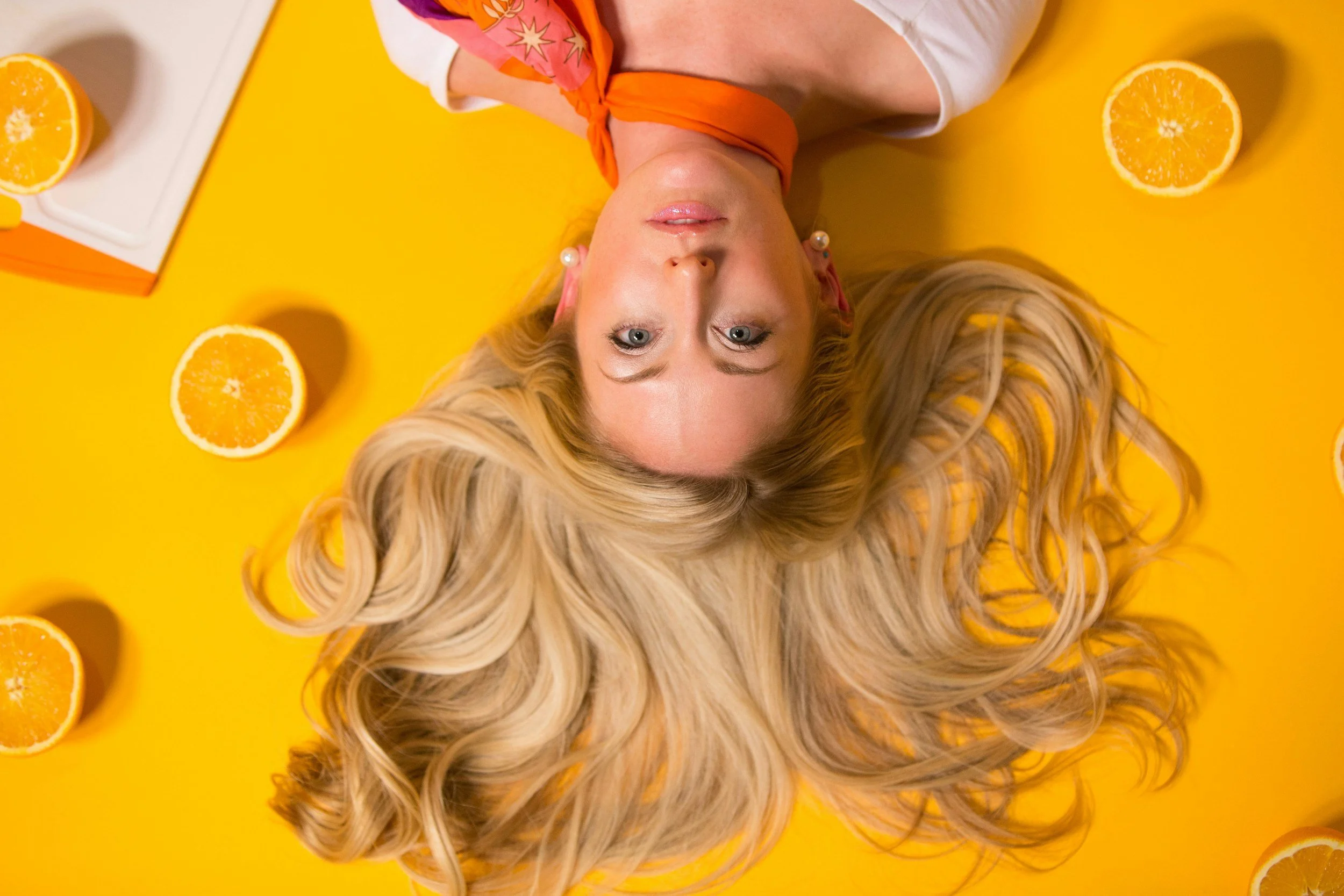 A woman with long, blonde, wavy hair lying on a yellow surface, surrounded by halved oranges, with her head positioned upside down, wearing a colorful outfit.