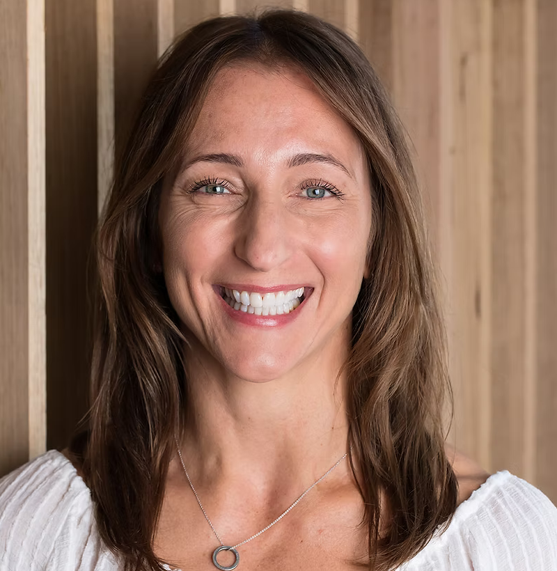 A woman smiling with light blue eyes and brown wavy hair, wearing a white top and a silver necklace, standing against a wooden background.