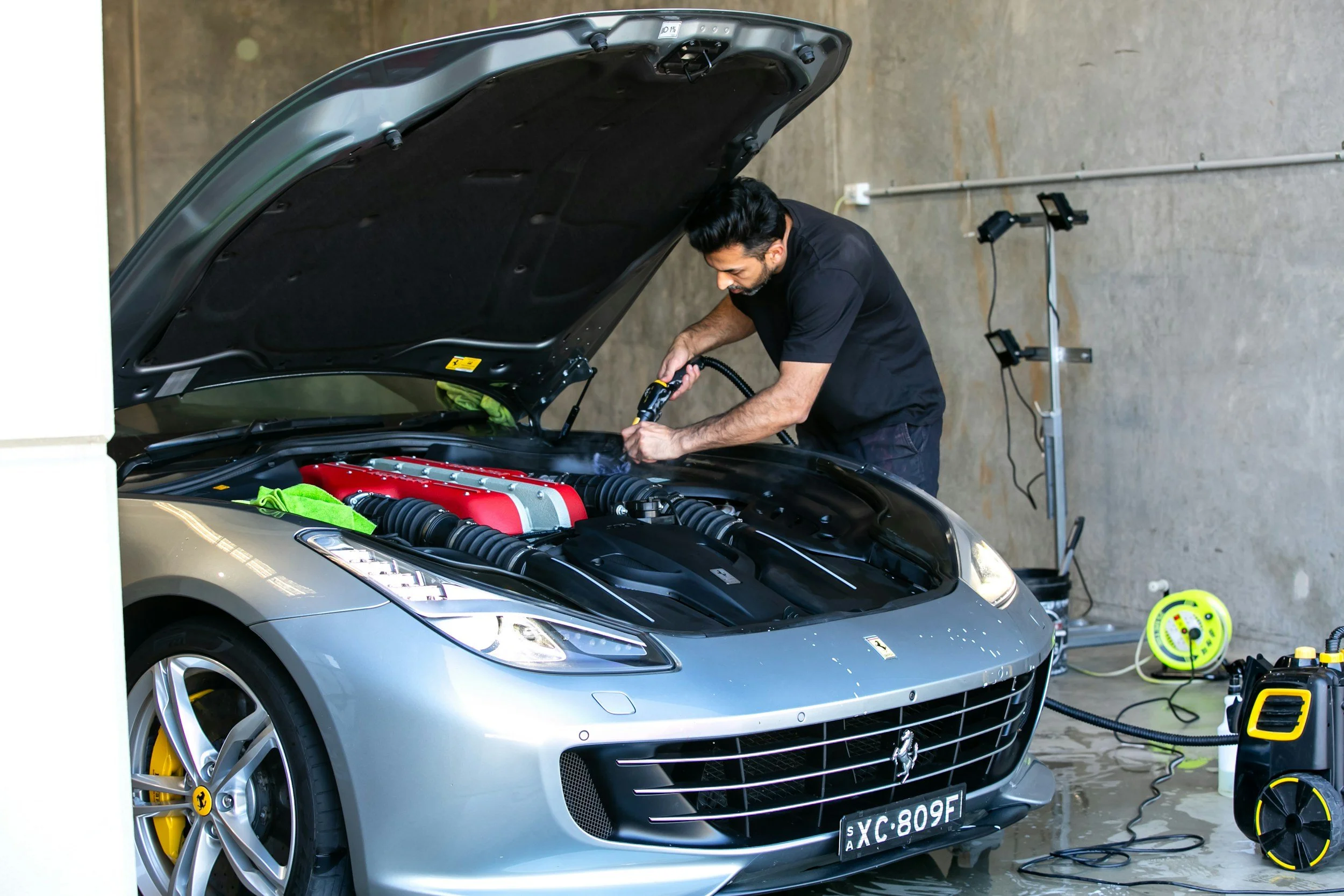 A man washing a silver Ferrari sports car inside a garage, connected to cleaning and detailing equipment.