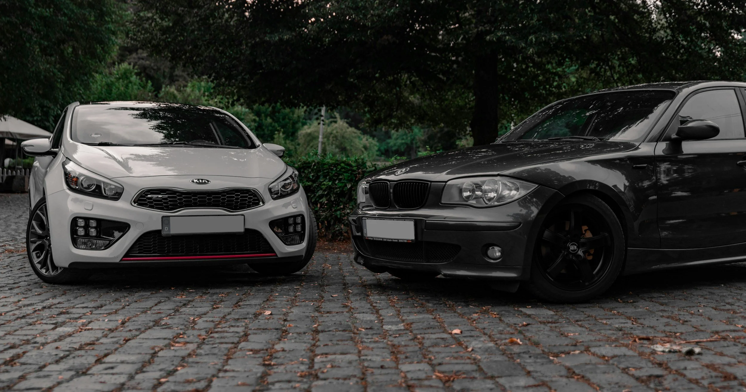 A white Kia and a black BMW parked facing each other on a cobblestone street, with trees and greenery in the background.