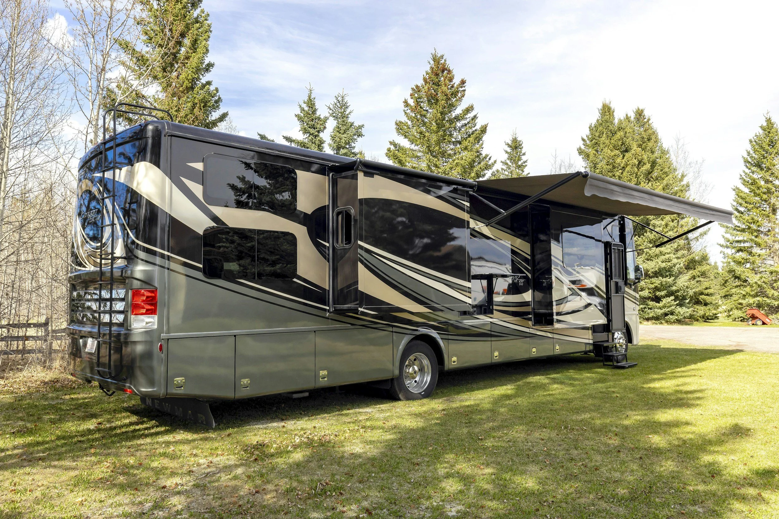 A large black and gray recreational vehicle with beige and black stripe designs, parked on a grassy area with trees in the background, and an extended retractable awning.