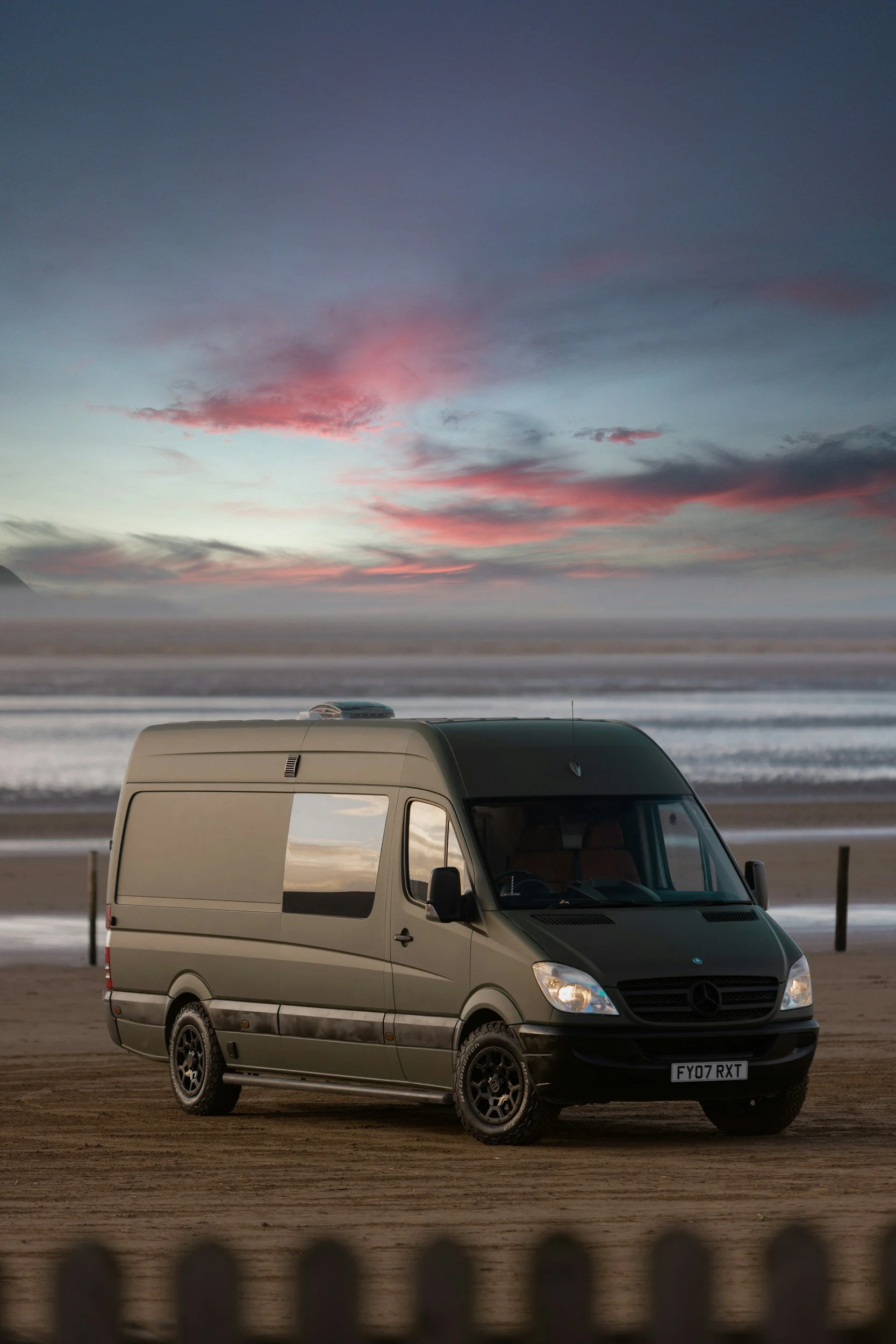 A tan Mercedes-Benz Sprinter van parked on a sandy beach at sunset, with the ocean in the background and a pink and blue sky.