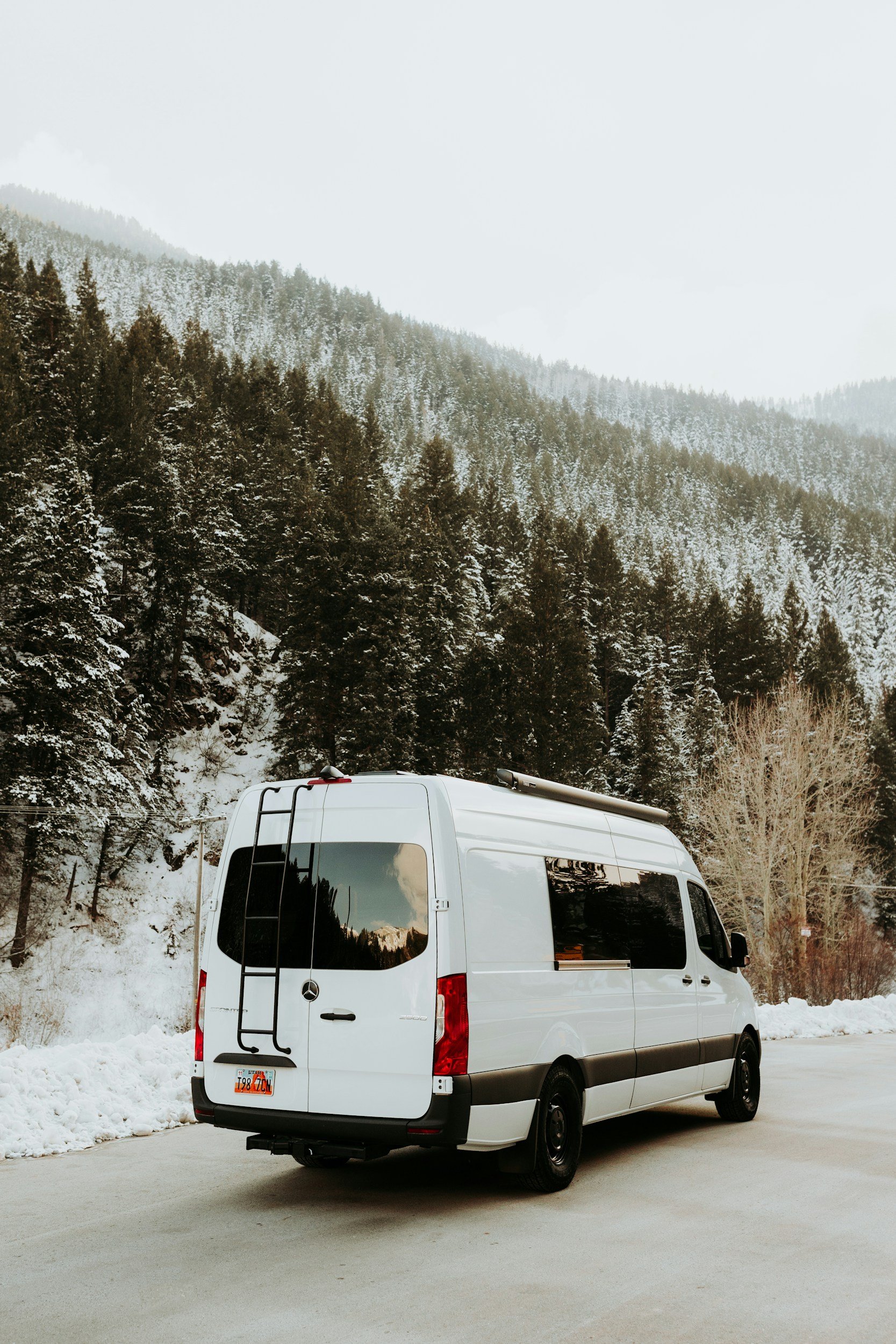 White van parked on a snow-covered roadside with a mountainous, forested background and snow on the trees.