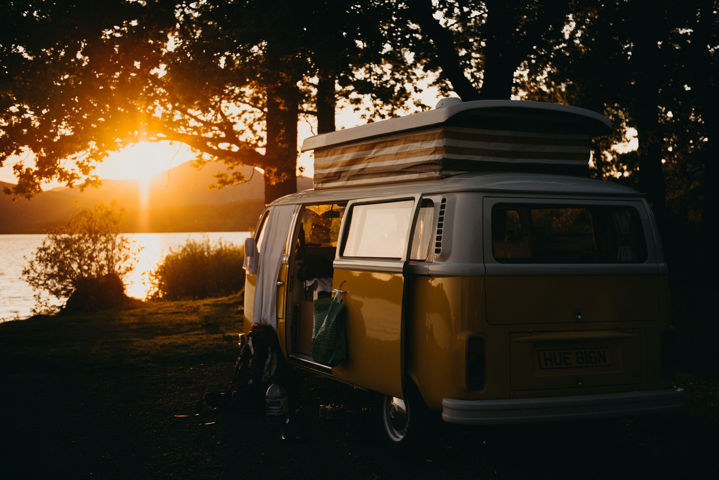Yellow vintage camper van parked near a lake at sunset, with a roof tent expanded on top, surrounded by trees.
