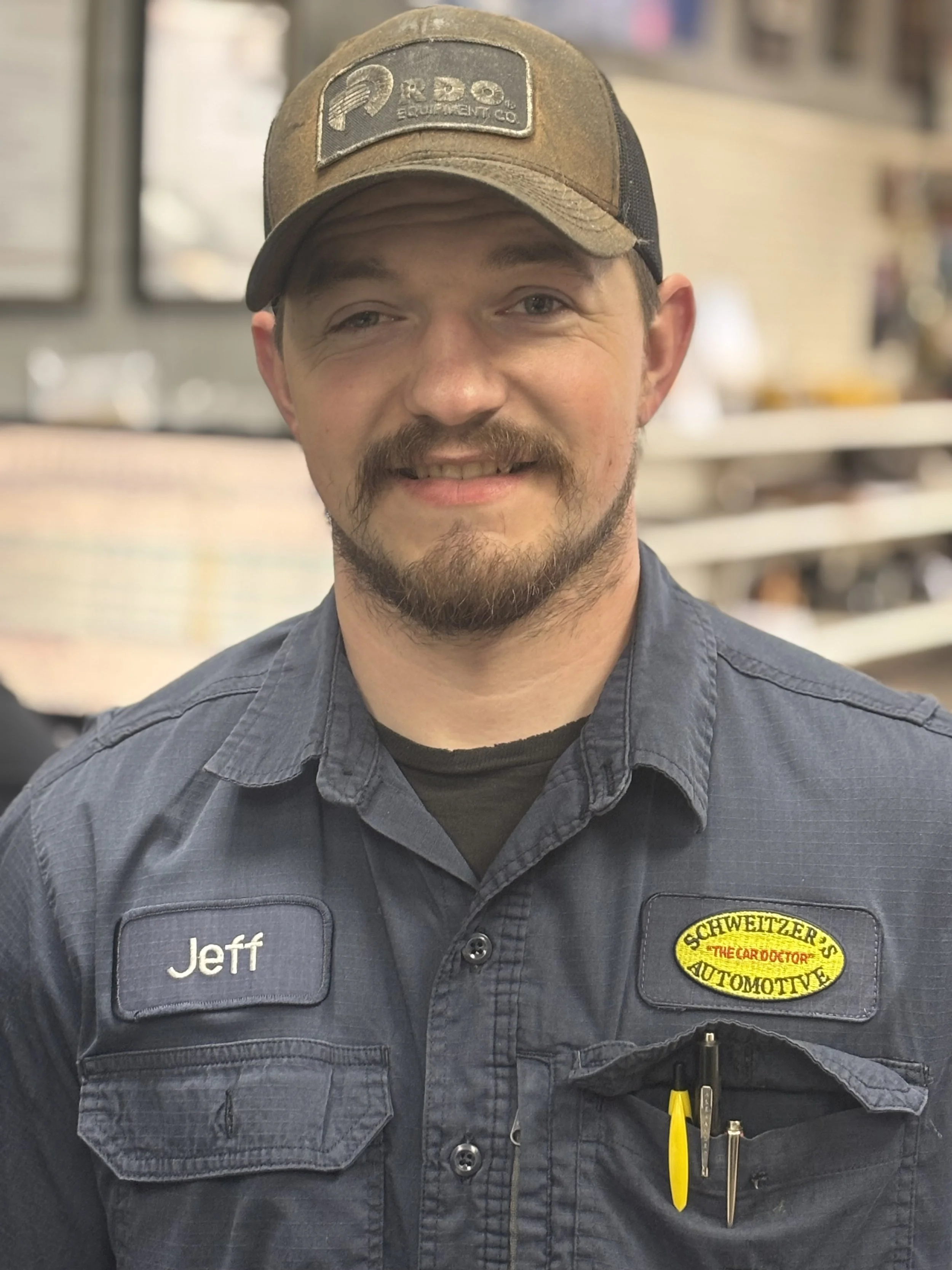 A man wearing a gray work shirt with a name tag that says 'Jeff' and a patch with the name 'Schweitzer Automotive.' He is also wearing a brown and black baseball cap with a logo. The background appears to be an auto repair shop or similar setting.