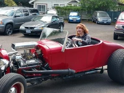 A red vintage-style convertible car with a woman driving, parked in a lot with modern cars and a building in the background.