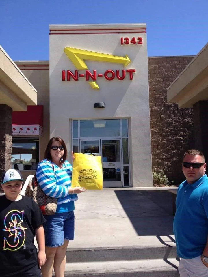 Three people standing in front of an In-N-Out Burger with a yellow bag. The building has a sign with a yellow arrow pointing towards the entrance, and the address 1342. The sky is clear and blue.