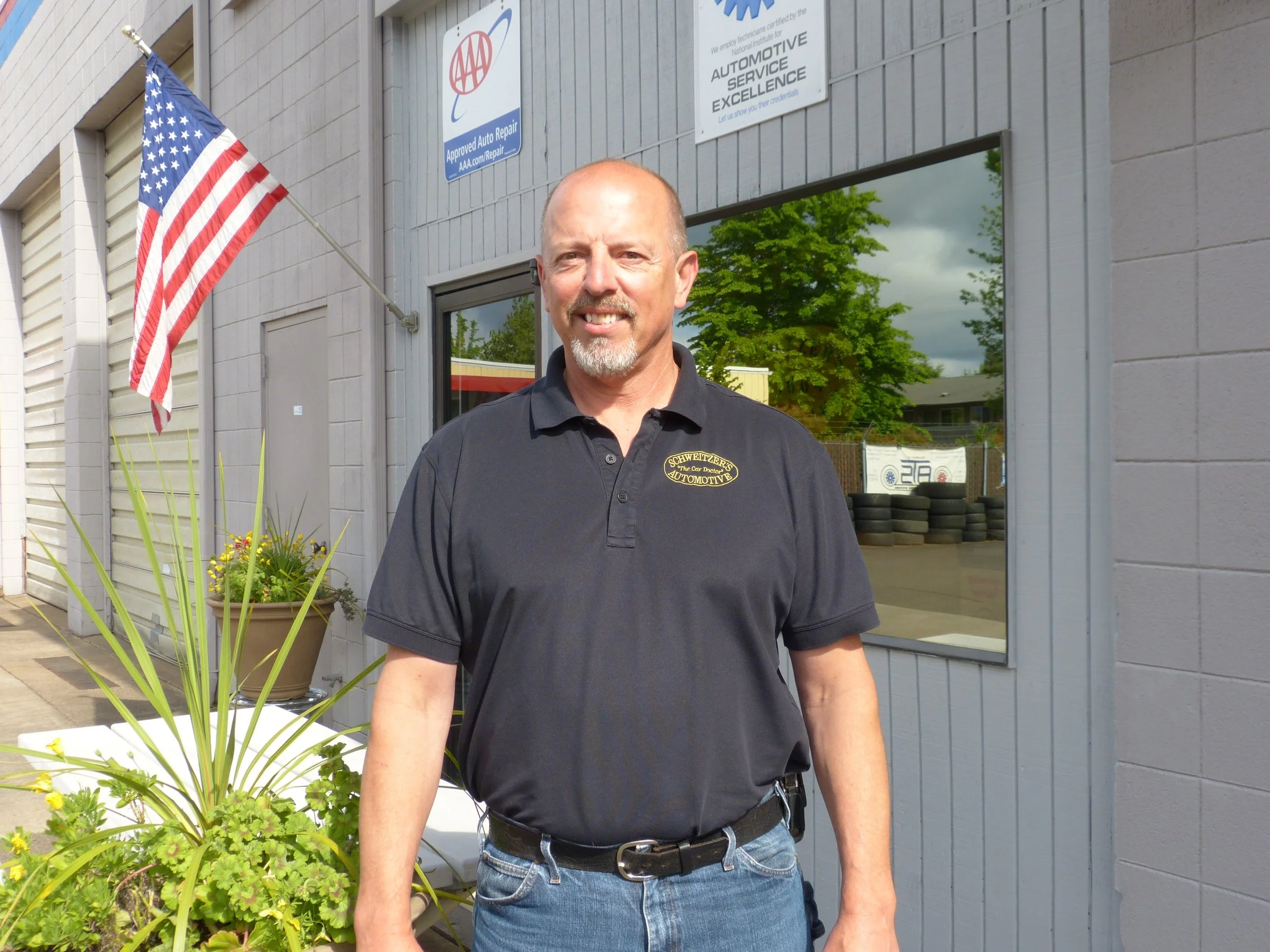 A man standing outside a building with a U.S. flag, reflective window showing trees, and signs for automotive service and AAA approval.