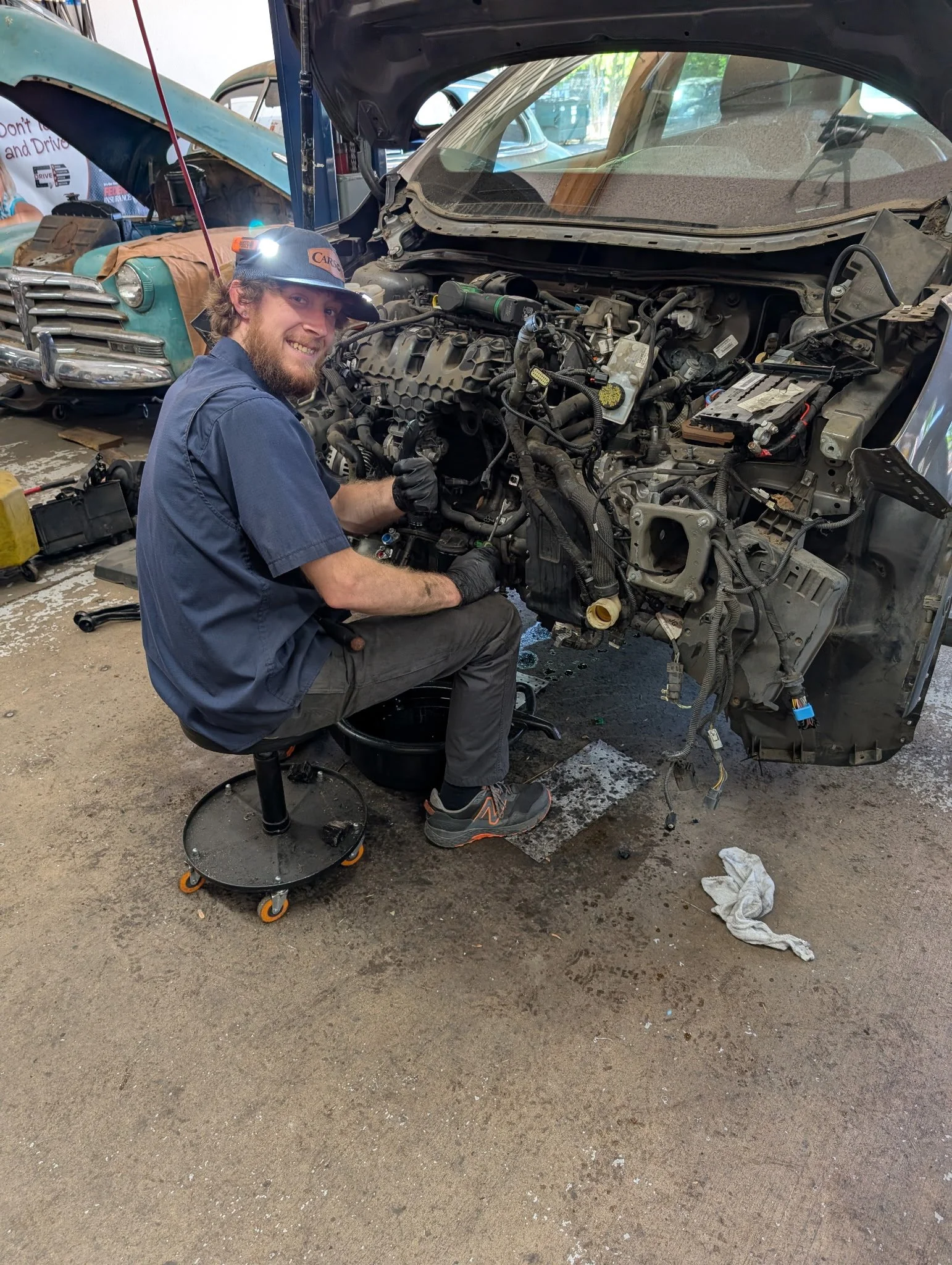 A mechanic working on a car engine in a garage, with a vintage car in the background.