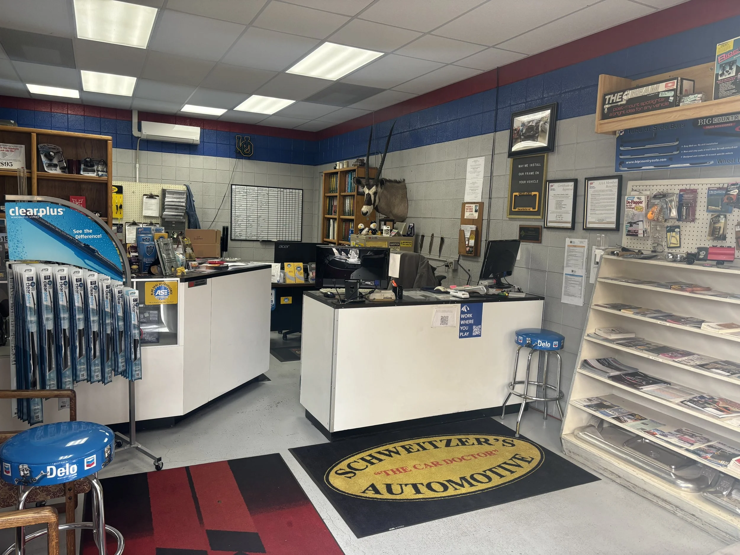 Interior of an automotive repair shop with a reception desk, shelves with tools and car accessories, a mounted animal head, and a branded mat on the floor reading 'Schweitzer's Automotive'.