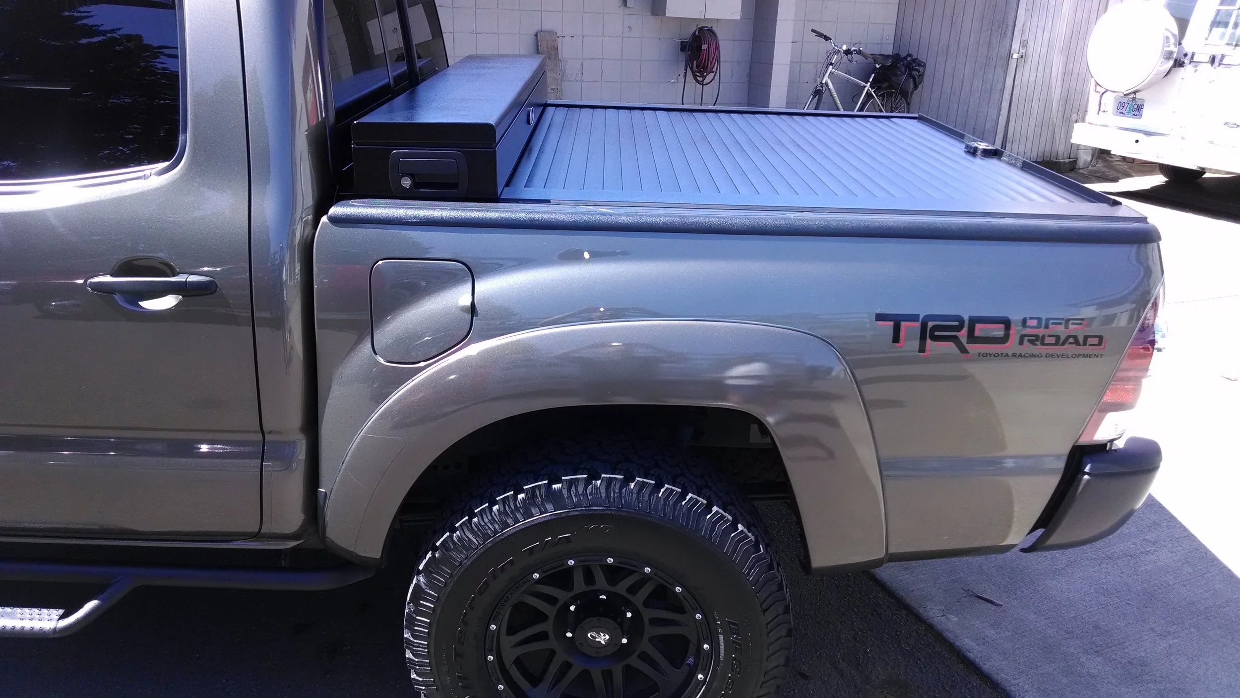 A silver Toyota pickup truck with a TRD Off Road decal on the rear side panel. The truck has a black tonneau cover over the bed and large off-road tires. There is a black box on the bed near the cab.