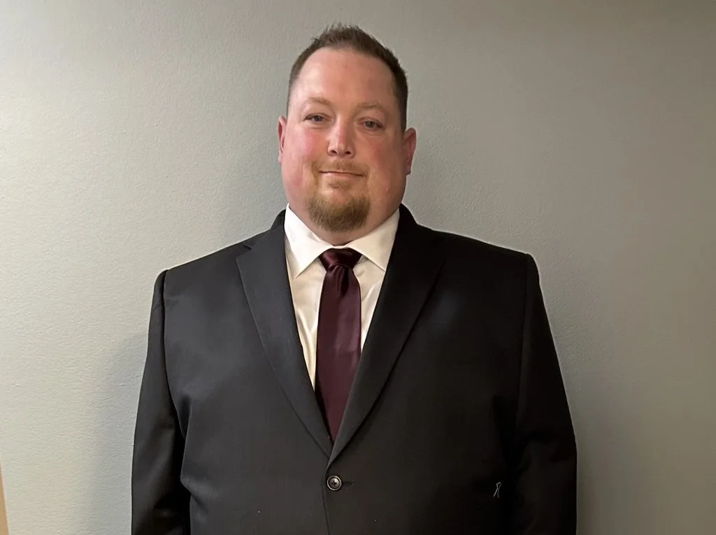 A man in a black suit, white shirt, and maroon tie standing against a plain gray wall.