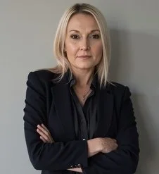 A woman with blonde hair wearing a black blazer, crossing her arms, standing against a gray wall