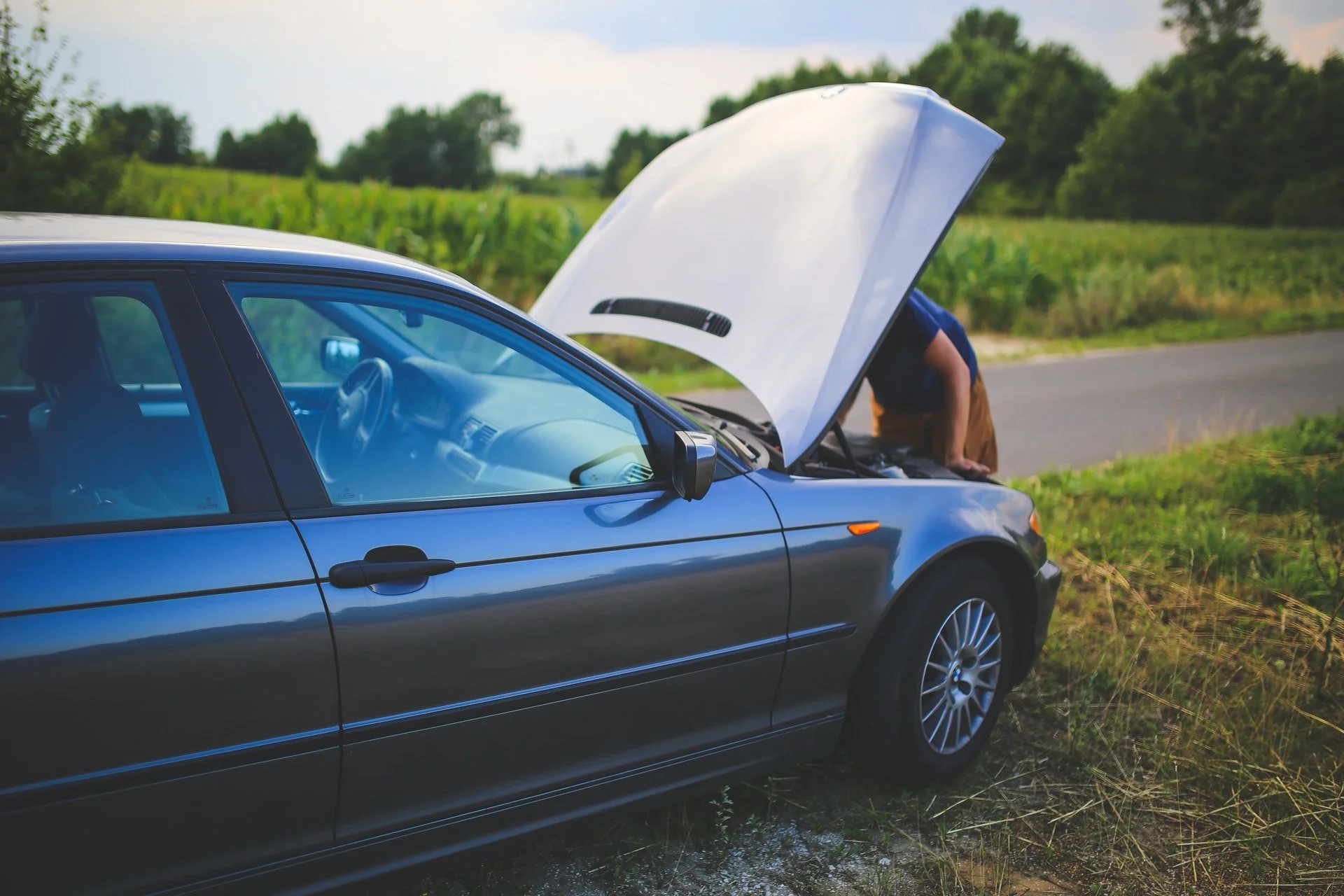 A person working on a blue car with its hood open on the side of a rural road surrounded by fields and trees.