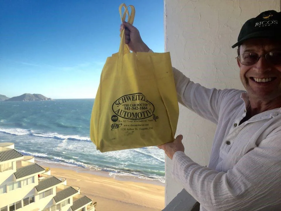 A man smiling on a balcony holds a yellow bag with 'Schweitzer Automotive' printed on it, with a beach and ocean in the background.