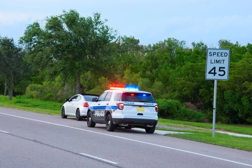 Police car with flashing lights pulled over a white sedan on the side of a rural road with a 45 mph speed limit sign, surrounded by green trees.