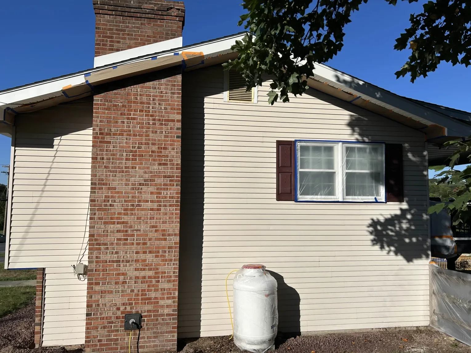 The side of a house with beige vinyl siding, a brick chimney, a window with brown shutters, blue painter's tape around the window frame, and a large white water heater outside. Part of a tree is visible at the top of the image.