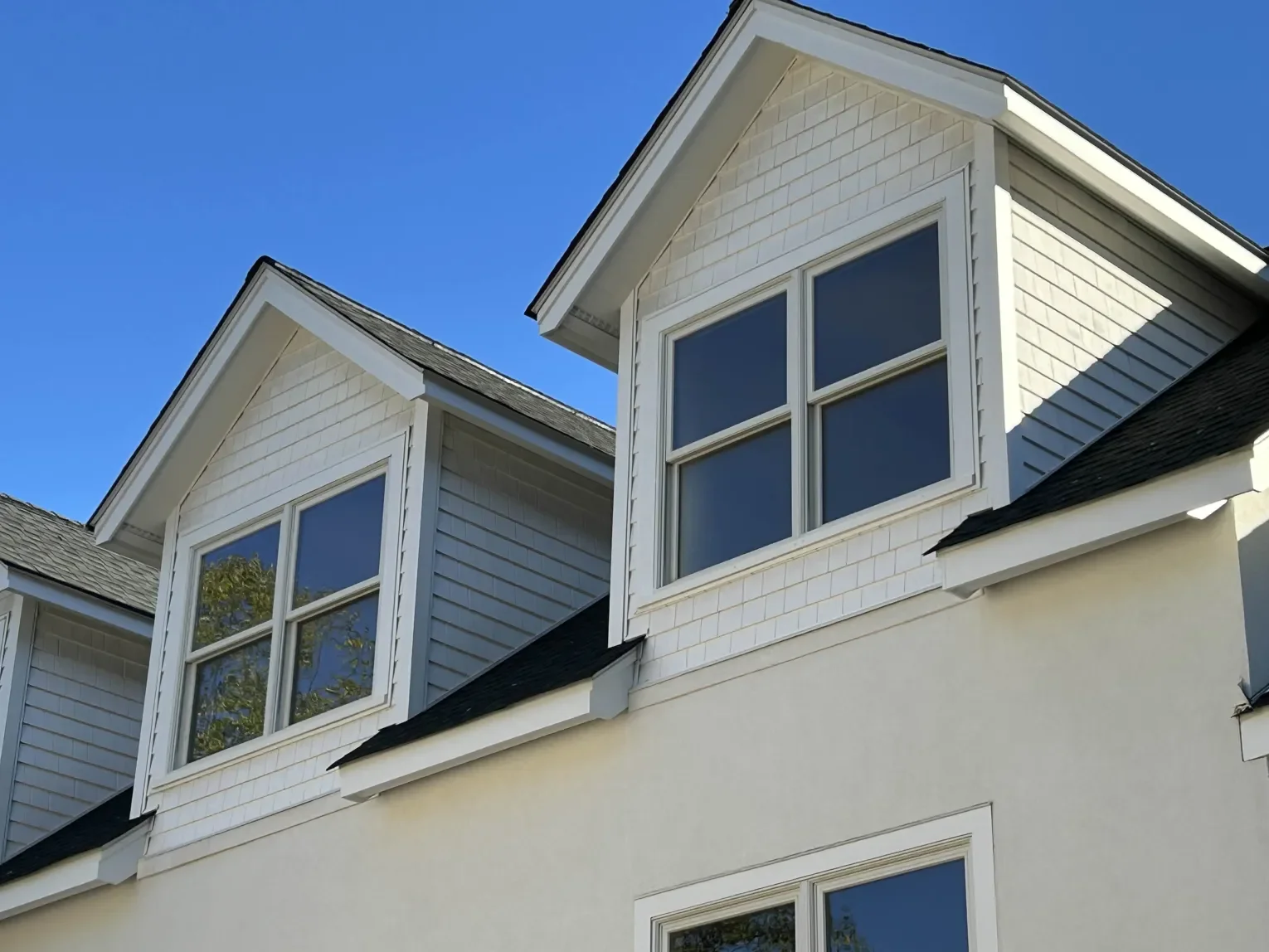 Close-up of a modern house facade showing white siding, large windows, and a black shingle roof, against a bright blue sky.